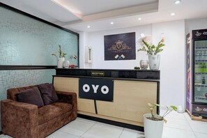 A reception area with a reception desk labeled 'OYO,' a couch, a wall-mounted fan, and a refrigerator stocked with beverages.
