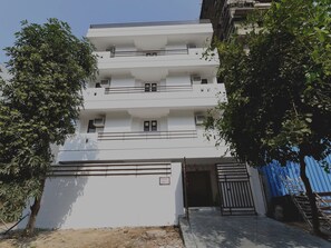 Um edifício residencial com vários andares, varandas, entrada com portão e uma cerca azul ao fundo.