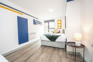 Habitación de hotel con cama, mesita de noche con lámpara, ventana y pared decorada con arte abstracto.
