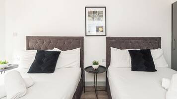 Ein Hotelzimmer mit zwei Betten, jeweils mit dunklem Kopfteil und weißer Bettwäsche. Zwischen den Betten hängt ein gerahmtes Bild an der Wand, und auf dem Boden steht ein kleiner Tisch mit einer Pflanze und einer Fernbedienung.