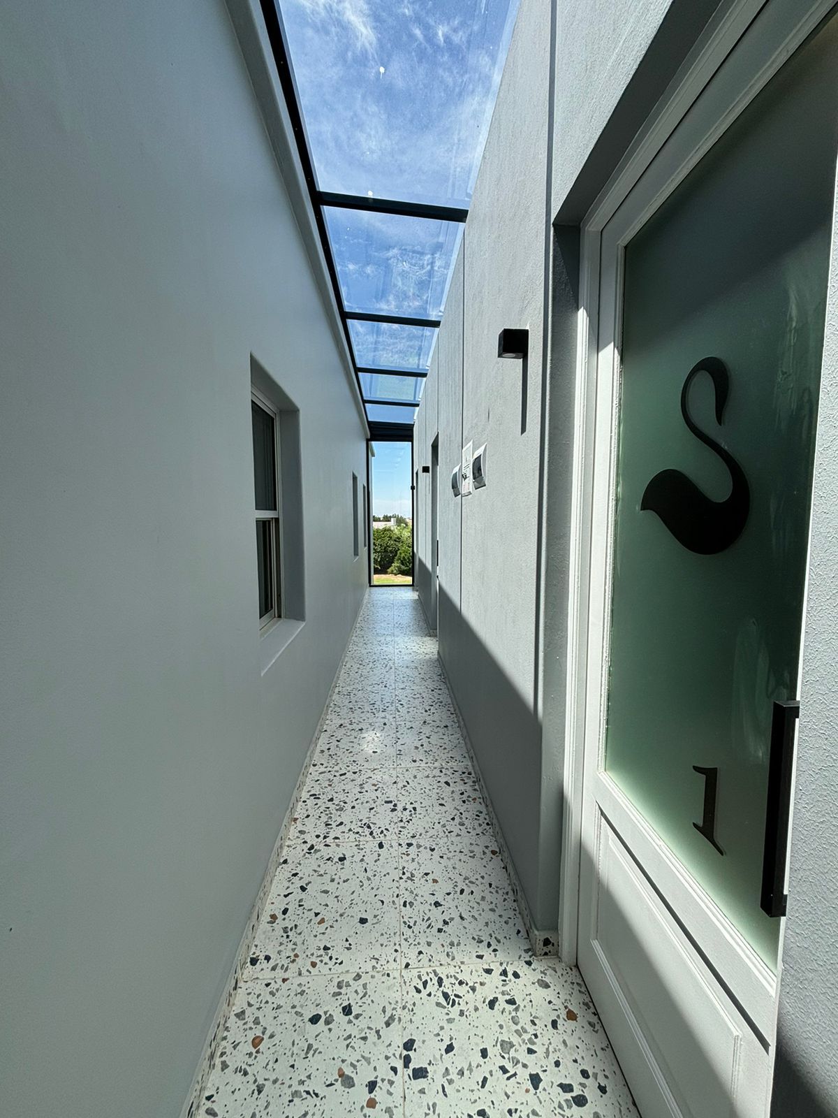 Ein Korridor mit Terrazzo-Boden, einer Glastür mit dem Buchstaben 'S' und einem klaren Himmel darüber.