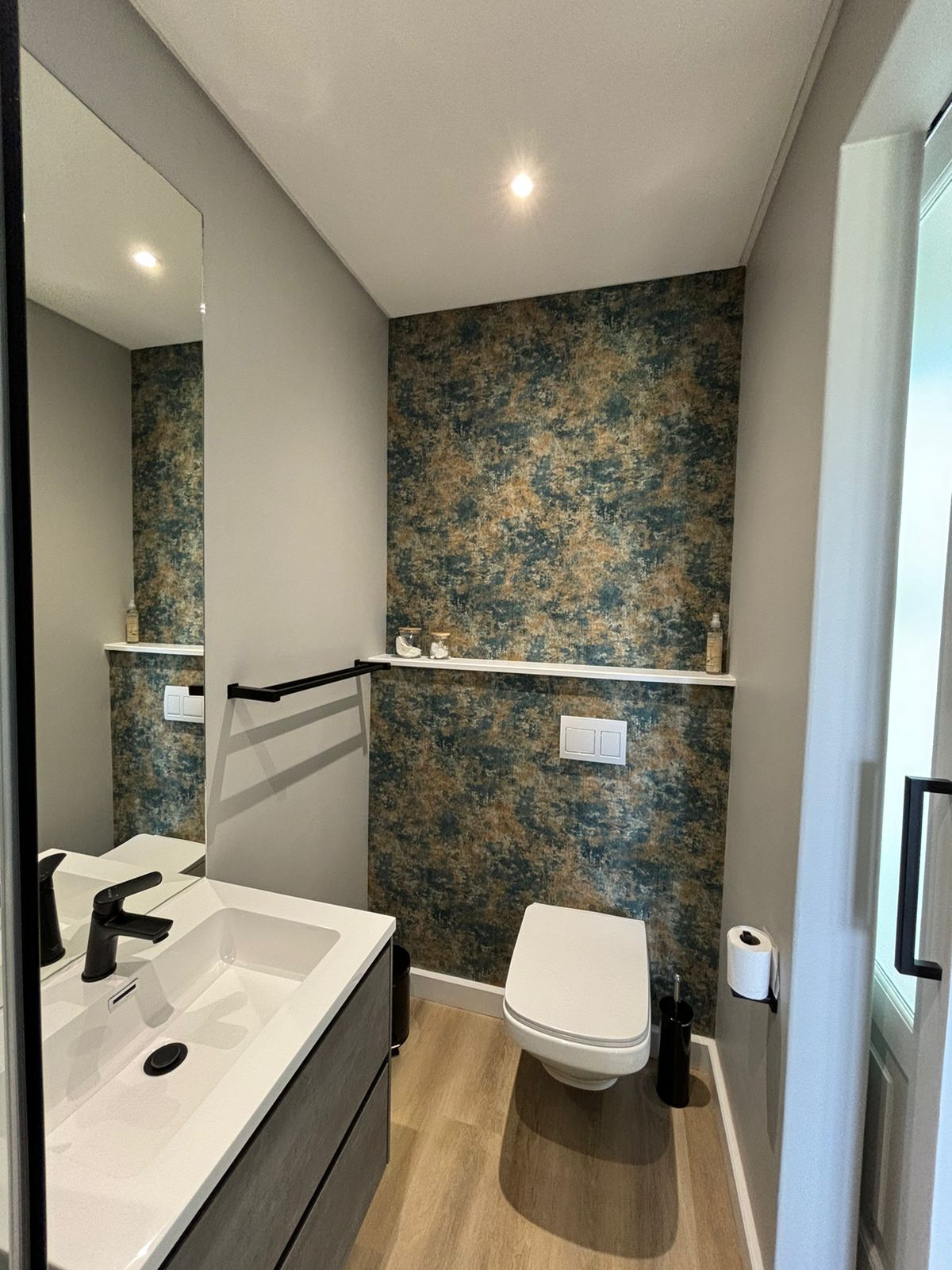 A bathroom with a white sink, a toilet, and a mirror. There are shelves above the toilet and a light switch on the wall.