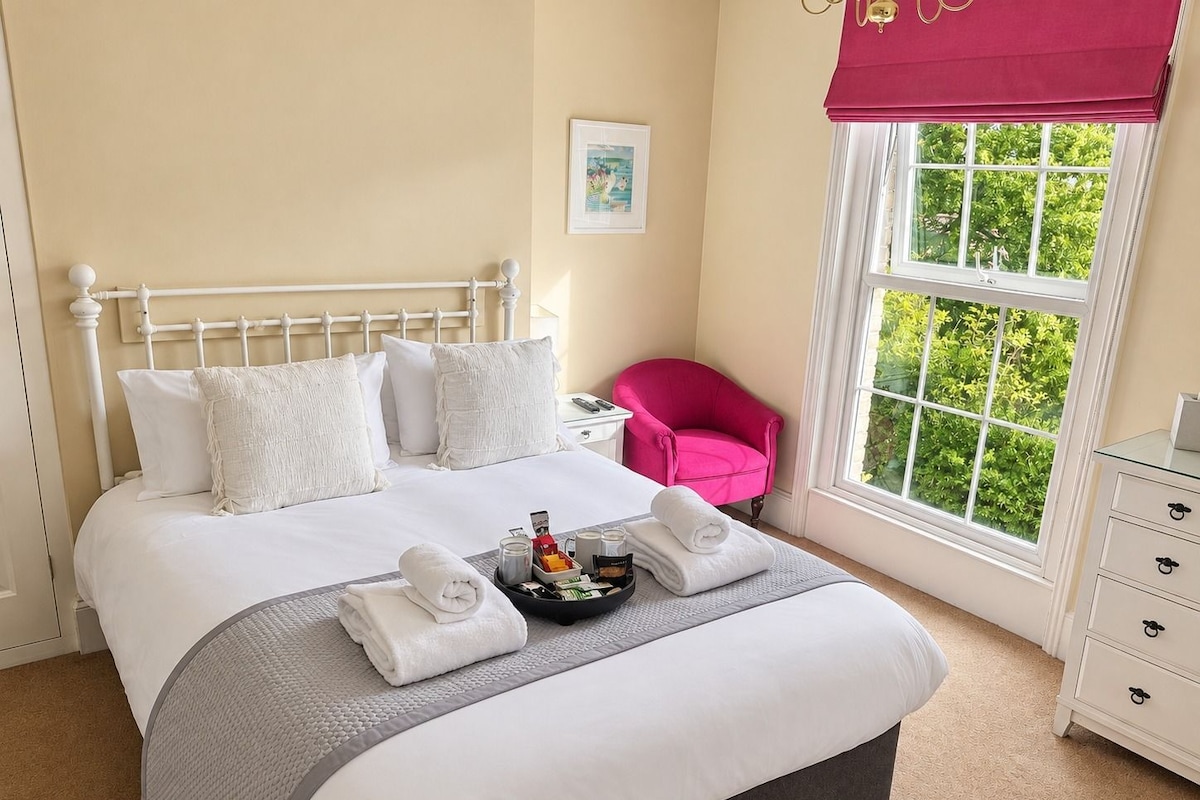 Una cama bien hecha con sábanas blancas y un detalle en gris, una silla rosa, una mesita de noche con una lámpara, y una ventana con vistas a zonas verdes.