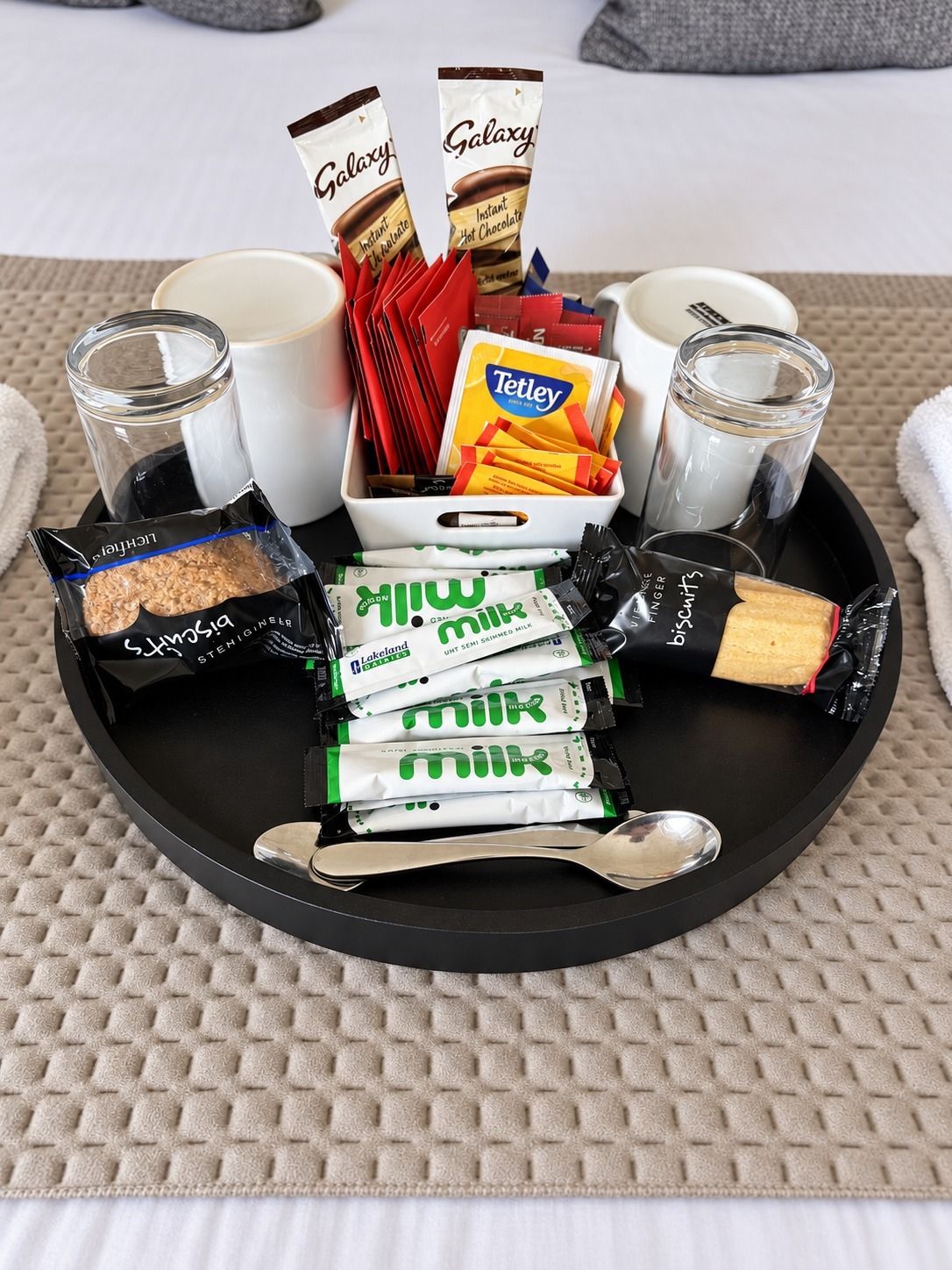 Bandeja com lanches, sachês de chocolate quente instantâneo, saquinhos de chá Tetley e um pote de leite.