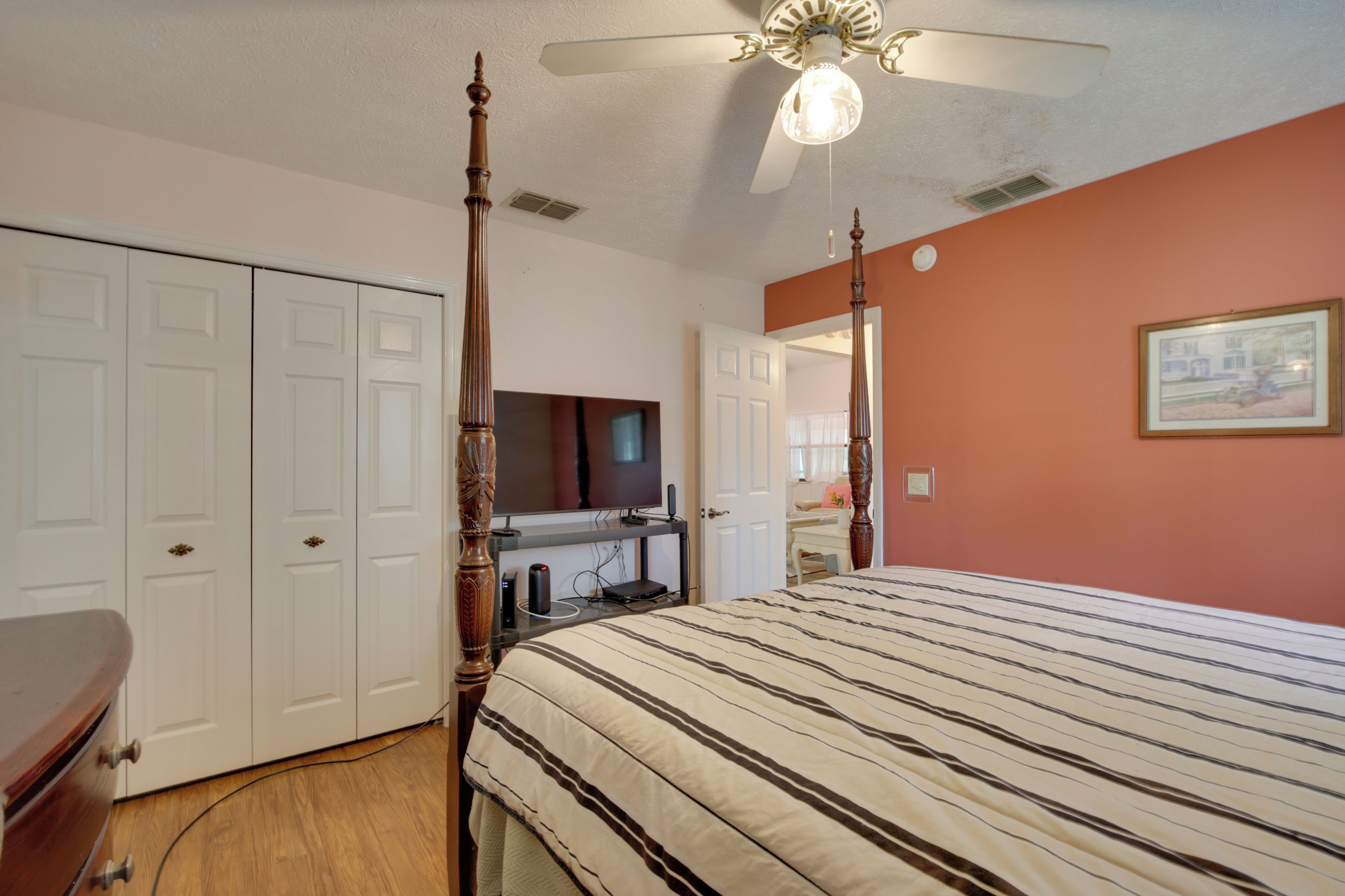 A bedroom with a four-poster bed, a ceiling fan, a television, and a door leading to another room.