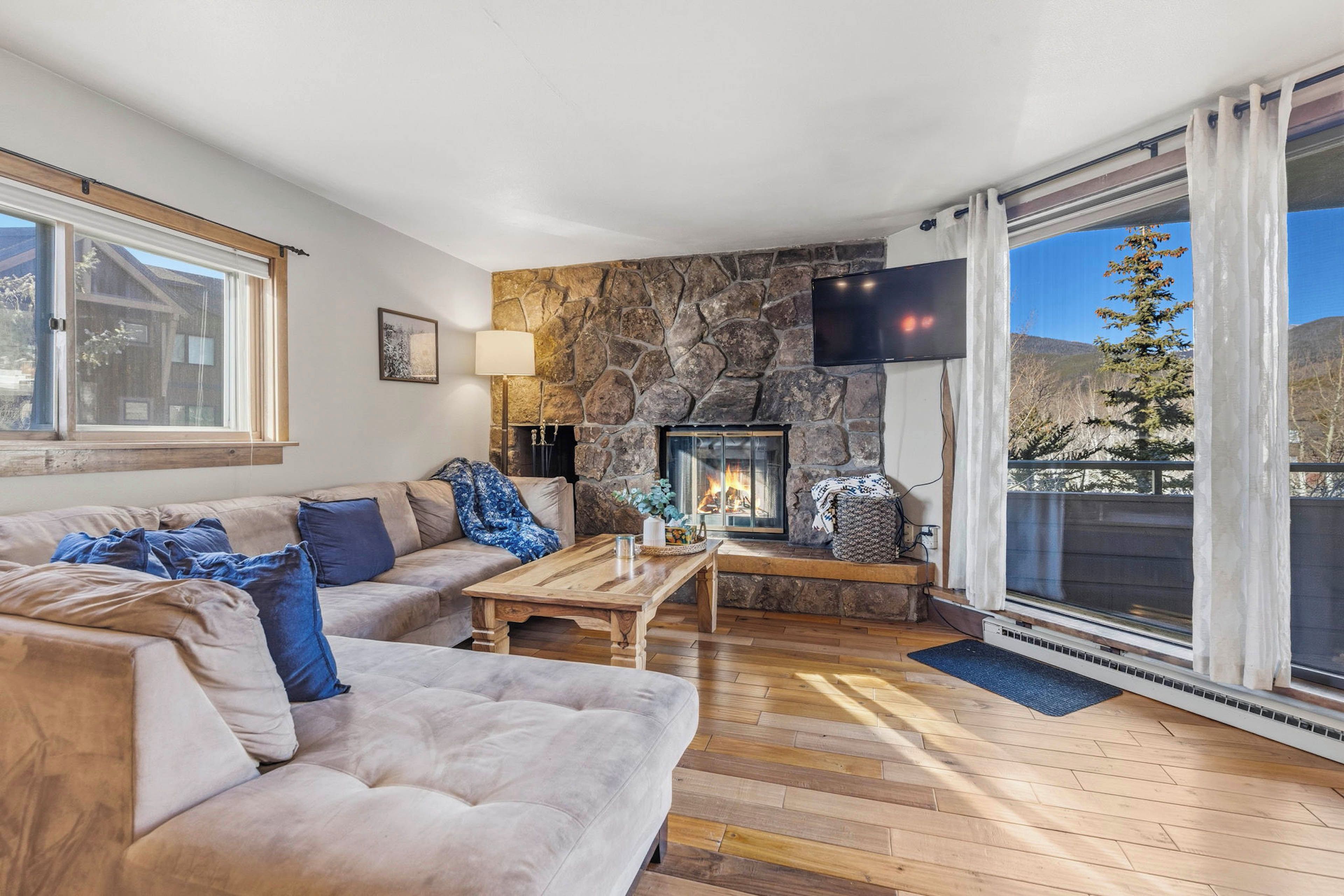 A living room with a stone fireplace, a flat-screen TV, and a large window with a view of trees and mountains.