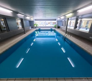 Piscina interna com pistas demarcadas, cercada por paredes com janelas e chão revestido com azulejos.