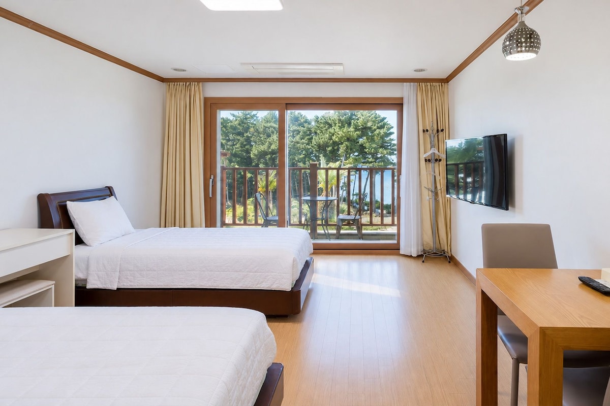 Ein Hotelzimmer mit zwei Betten, einem Schreibtisch, einem Stuhl, einem Fernseher und einem Balkon mit Blick auf Bäume und einen Pool.