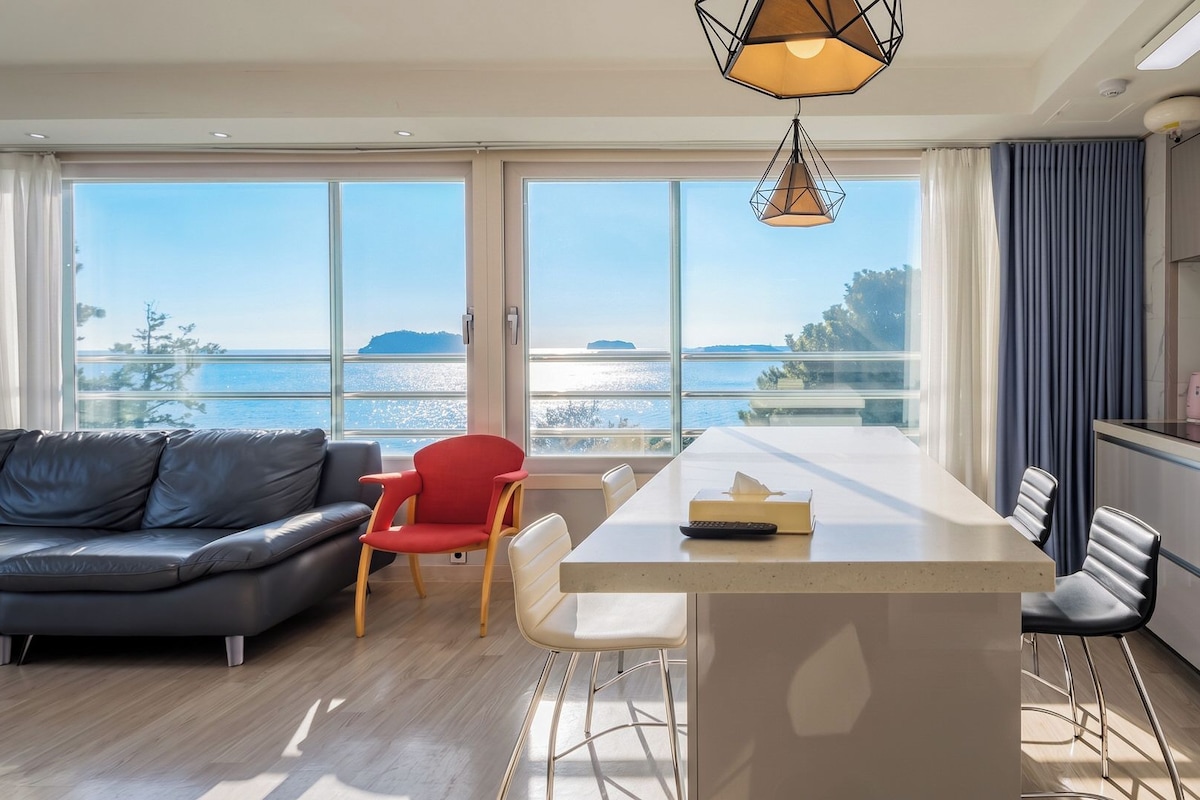 Ein modernes Wohnzimmer mit einer Couch, einem roten Sessel, einem Esstisch und einer Küchenzeile mit Meerblick.