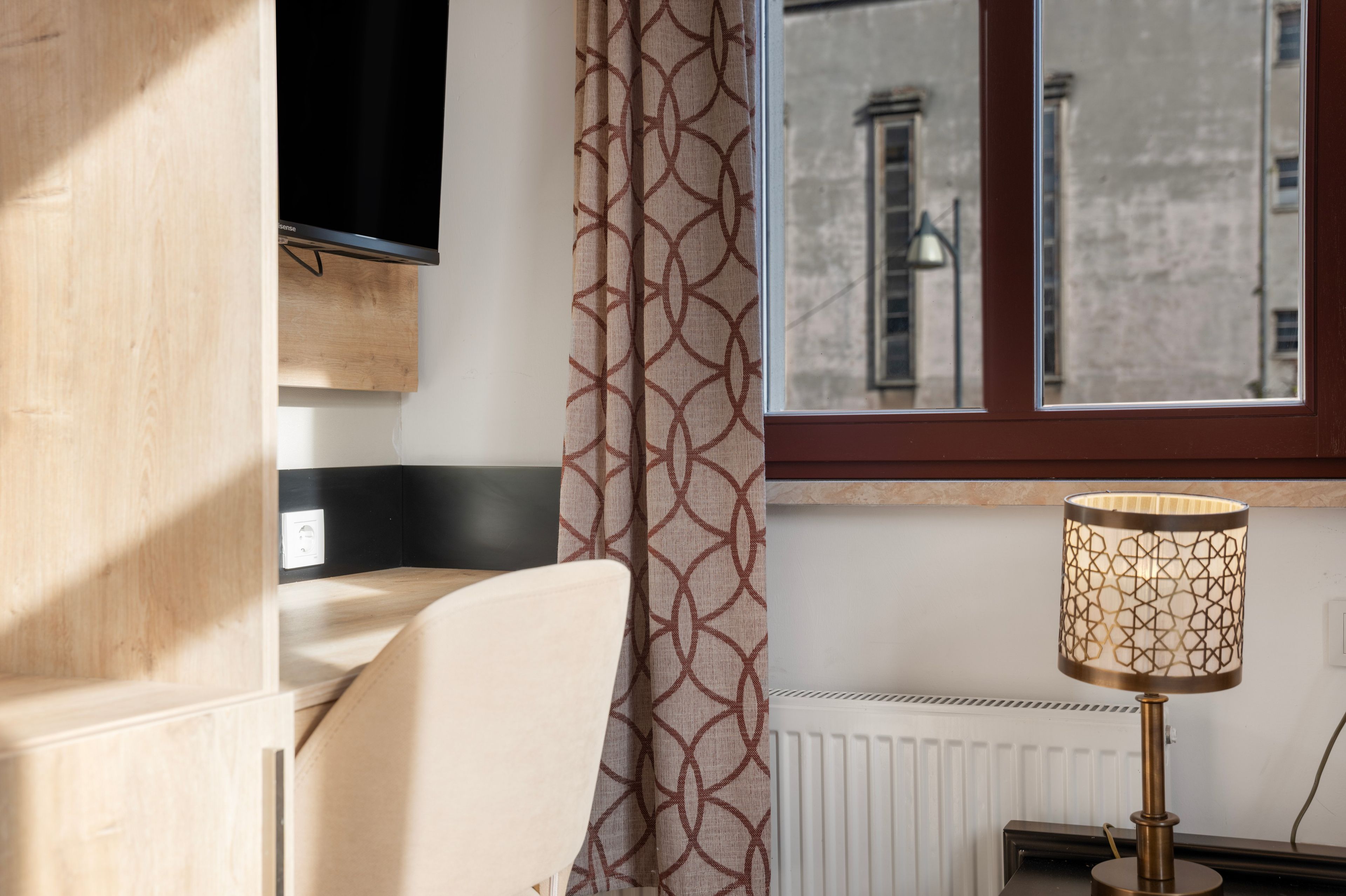 A room with a bed, a TV mounted on the wall, a patterned curtain, and a lamp on a table.