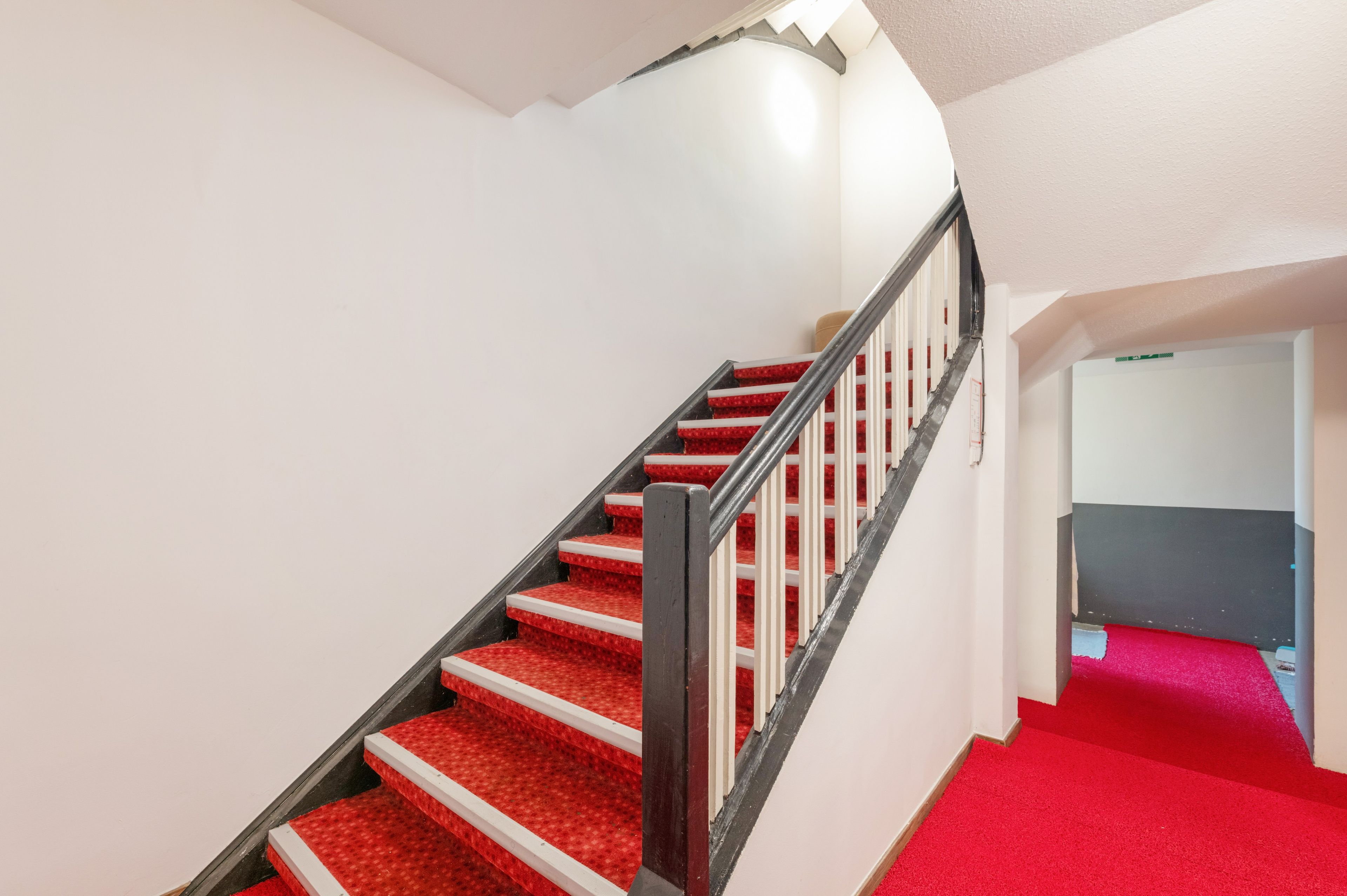 Eine Treppe mit roten Teppichstufen und einem schwarzen Geländer, die zu einem Raum mit weißer Wand und rotem Teppichboden führt.