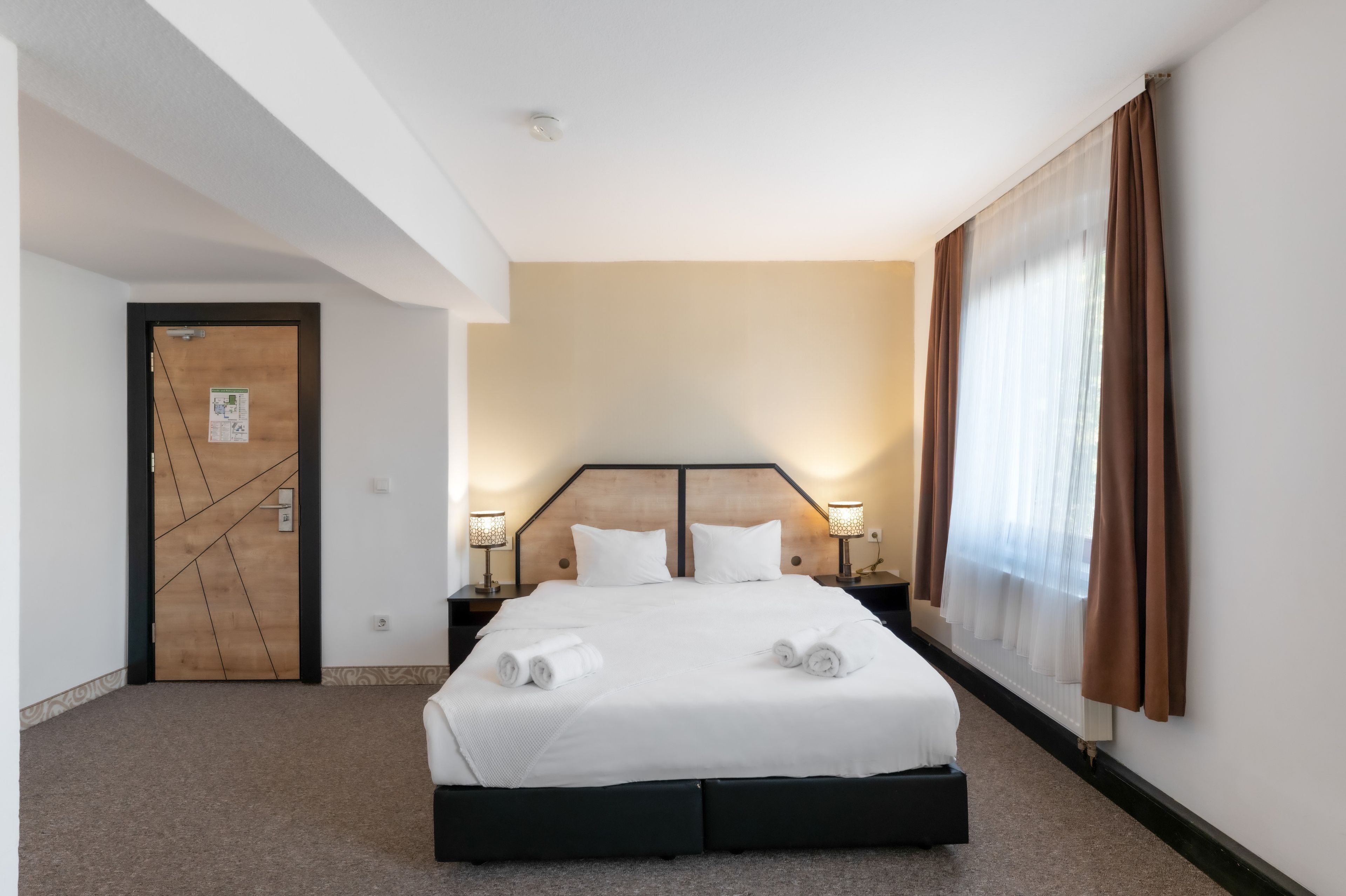 Ein Hotelzimmer mit einem Bett, zwei Nachttischlampen, einer Tür mit einer Karte und einem Fenster mit Vorhängen.