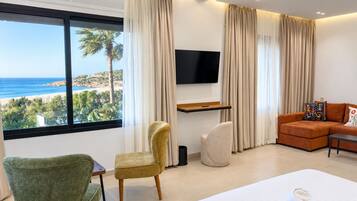 Habitación de hotel moderna con un gran ventanal que ofrece vistas a la playa y palmeras, una cama con toalla blanca, un sofá y un televisor montado en la pared.