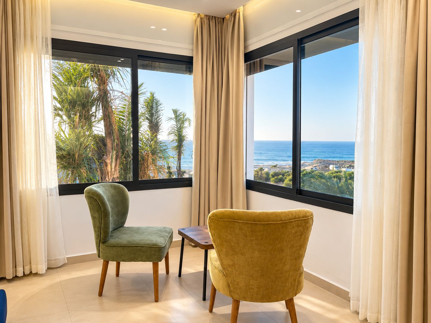 Ein Zimmer mit zwei Stühlen, einem kleinen Tisch und großen Fenstern mit Blick auf das Meer und Palmen.