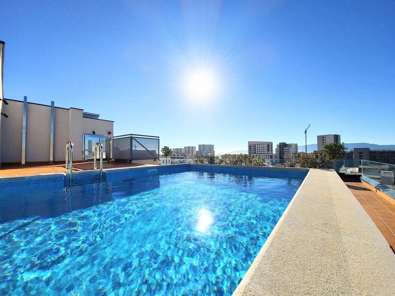 Una piscina en la azotea con agua cristalina y cielo despejado, rodeada de edificios.