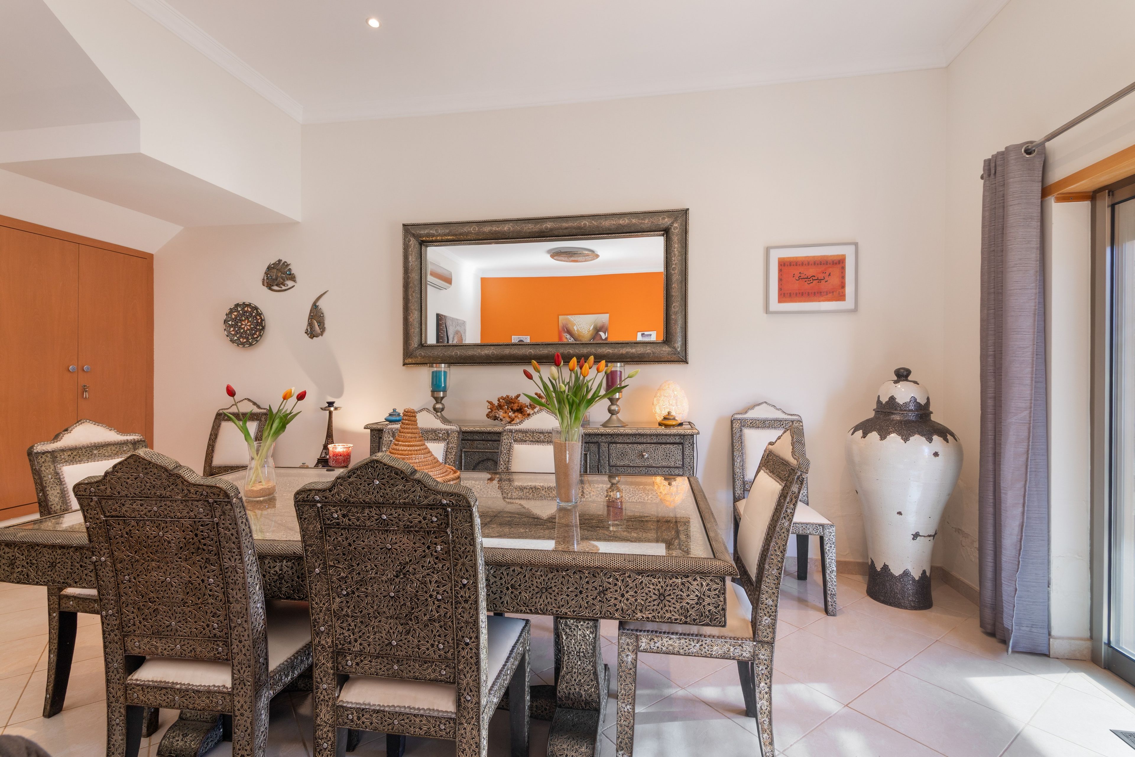 Un coin repas avec une table en verre entourée de chaises aux motifs complexes, un grand miroir au mur et des objets décoratifs, notamment un vase avec des fleurs et un chandelier.