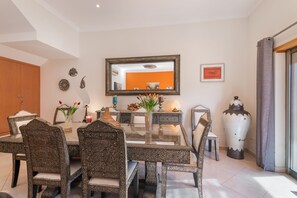 A dining area with a glass table surrounded by intricately patterned chairs, a large mirror on the wall, and decorative items including a vase with flowers and a candle holder. - Holiday Home 'Villa Quarto Crescente' with Shared Pool, Private Terrace and Air Conditioning (Albufeira)