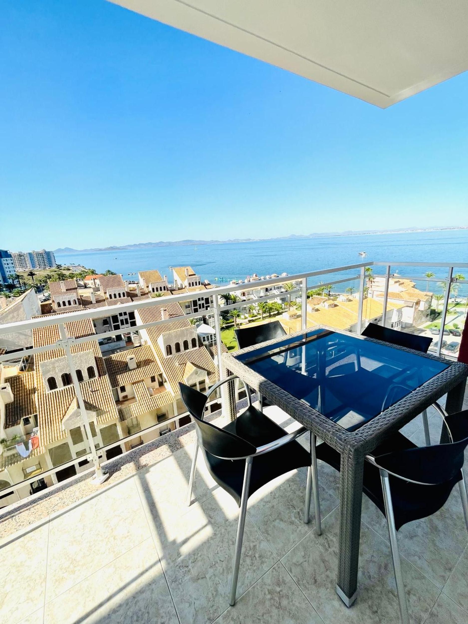 Un balcon doté d’une table et de chaises à plateau en verre, offrant une vue sur le paysage urbain de la côte, avec ses bâtiments et sa vue sur la mer.