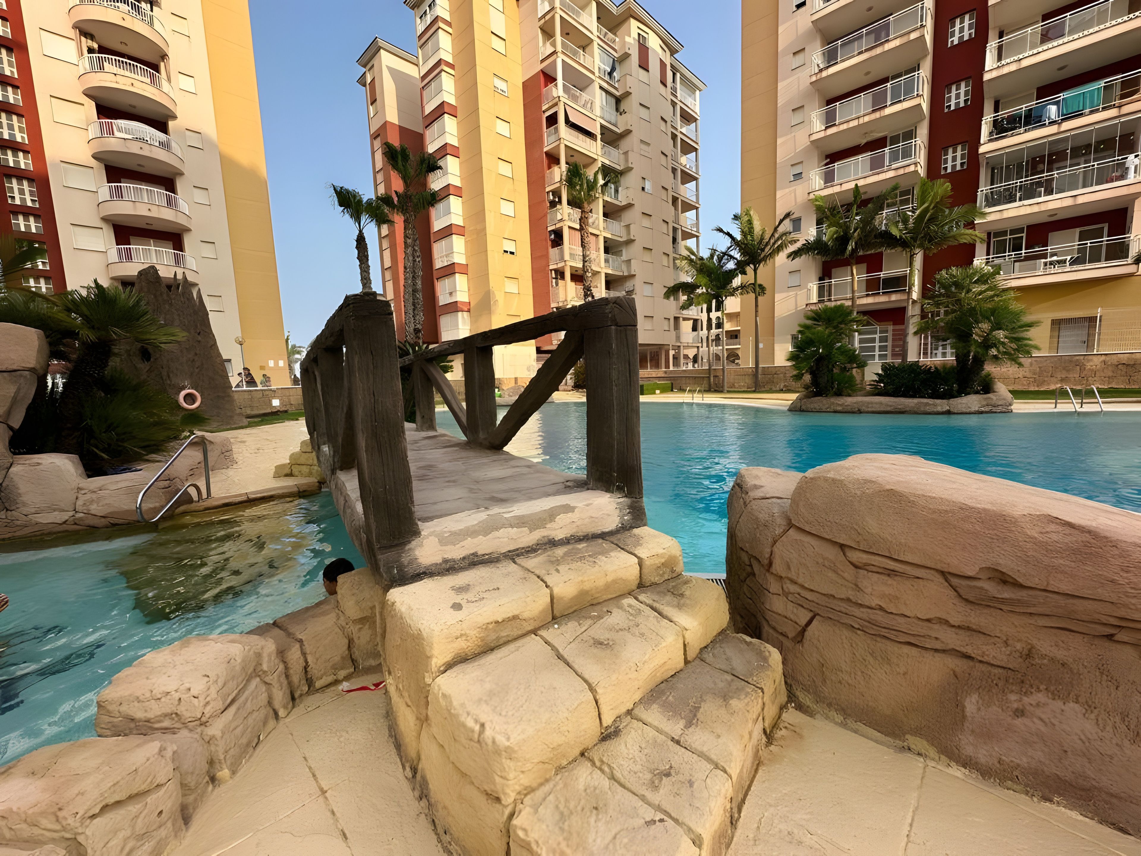 Ein Schwimmbecken mit einer Holztreppe, die zum Wasser führt, umgeben von Palmen und einem mehrstöckigen Gebäude mit Balkonen.