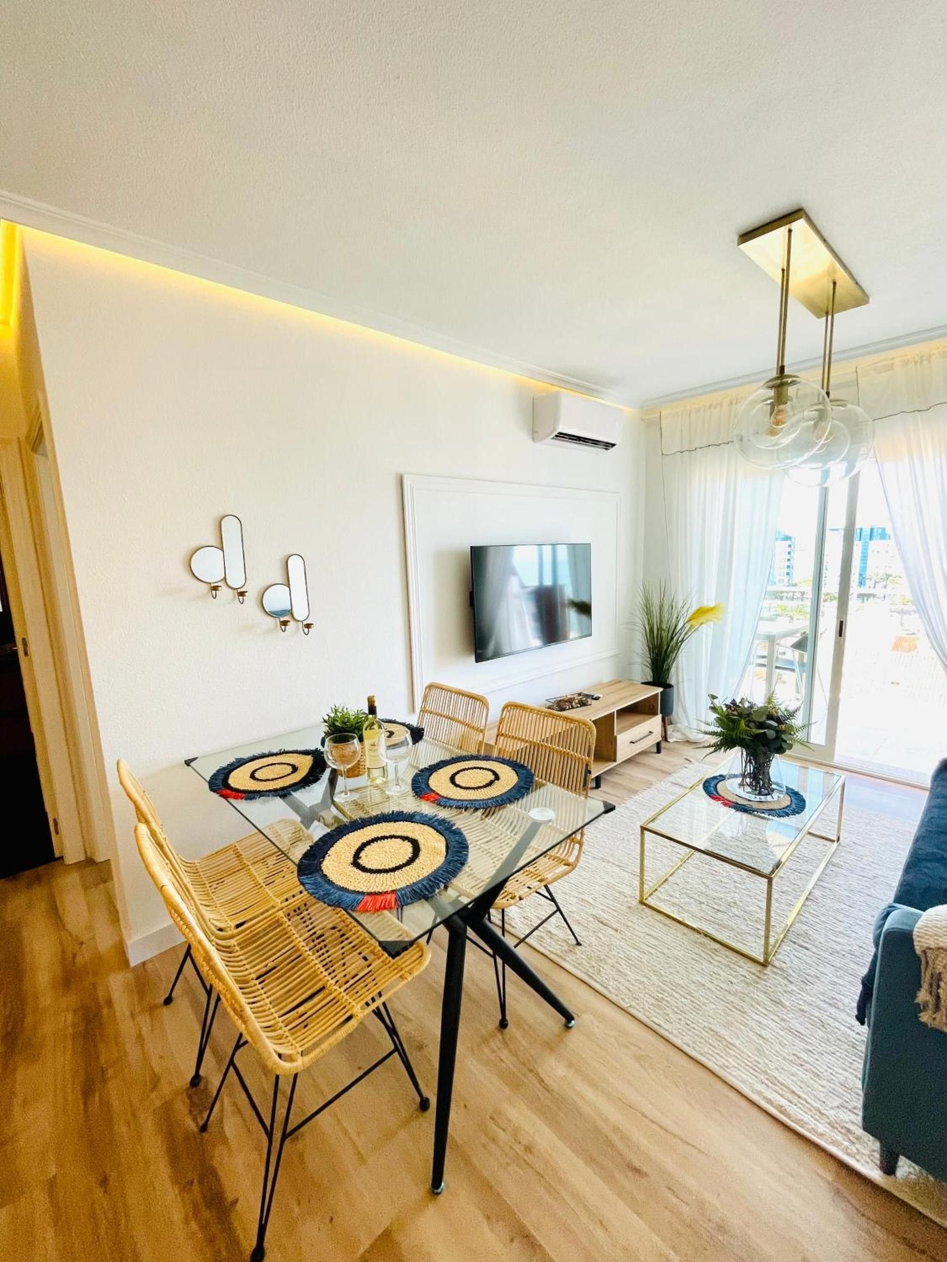 A modern living room with a glass dining table, wicker chairs, a flat-screen TV, and a sofa.