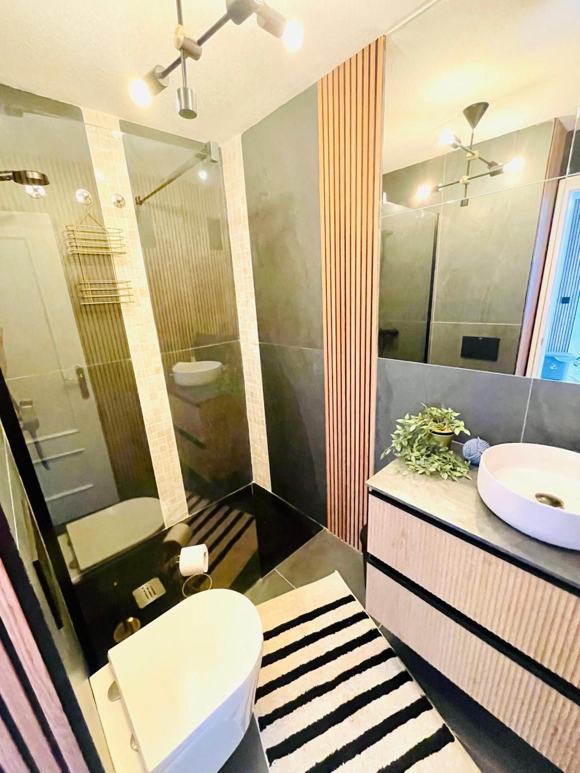 Un baño moderno con cabina de ducha de vidrio, inodoro blanco, alfombra a rayas negras y blancas, mueble de tocador de madera con lavamanos redondo y un espejo.