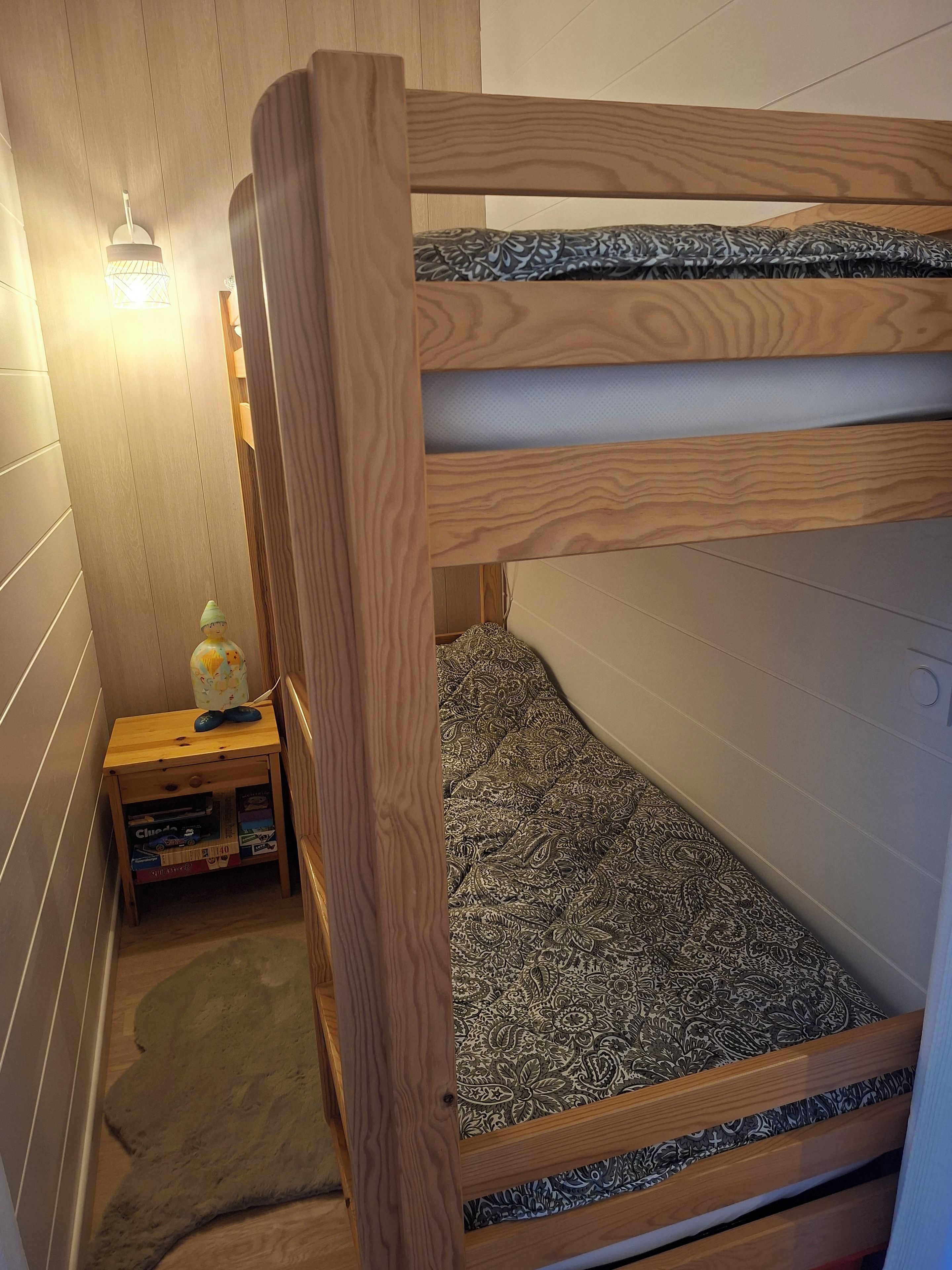 Un lit superposé avec un matelas à motifs, une petite table de chevet en bois sur laquelle se trouvent un globe terrestre et des livres, et un luminaire fixé au mur.