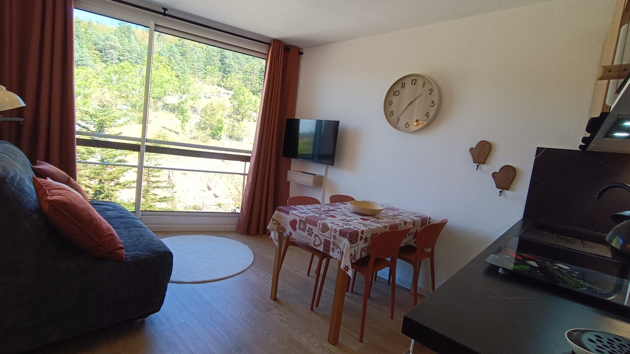 Un salon comprenant un canapé, une table à manger avec des chaises, un téléviseur à écran plat, une horloge et un coin cuisine avec une cuisinière.