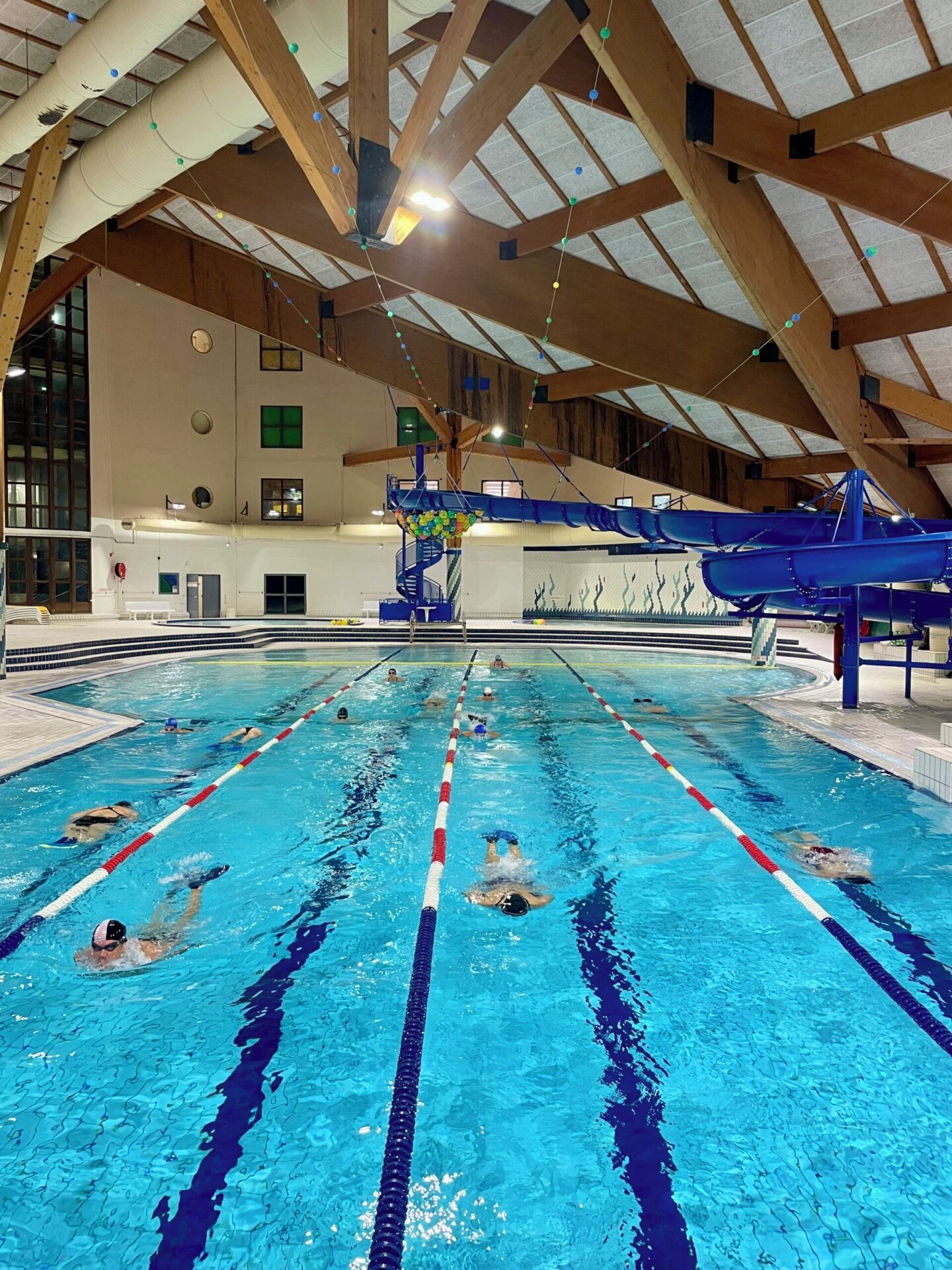 Une piscine intérieure avec des couloirs clairement délimités, un toboggan aquatique bleu et un plafond haut avec des poutres en bois.