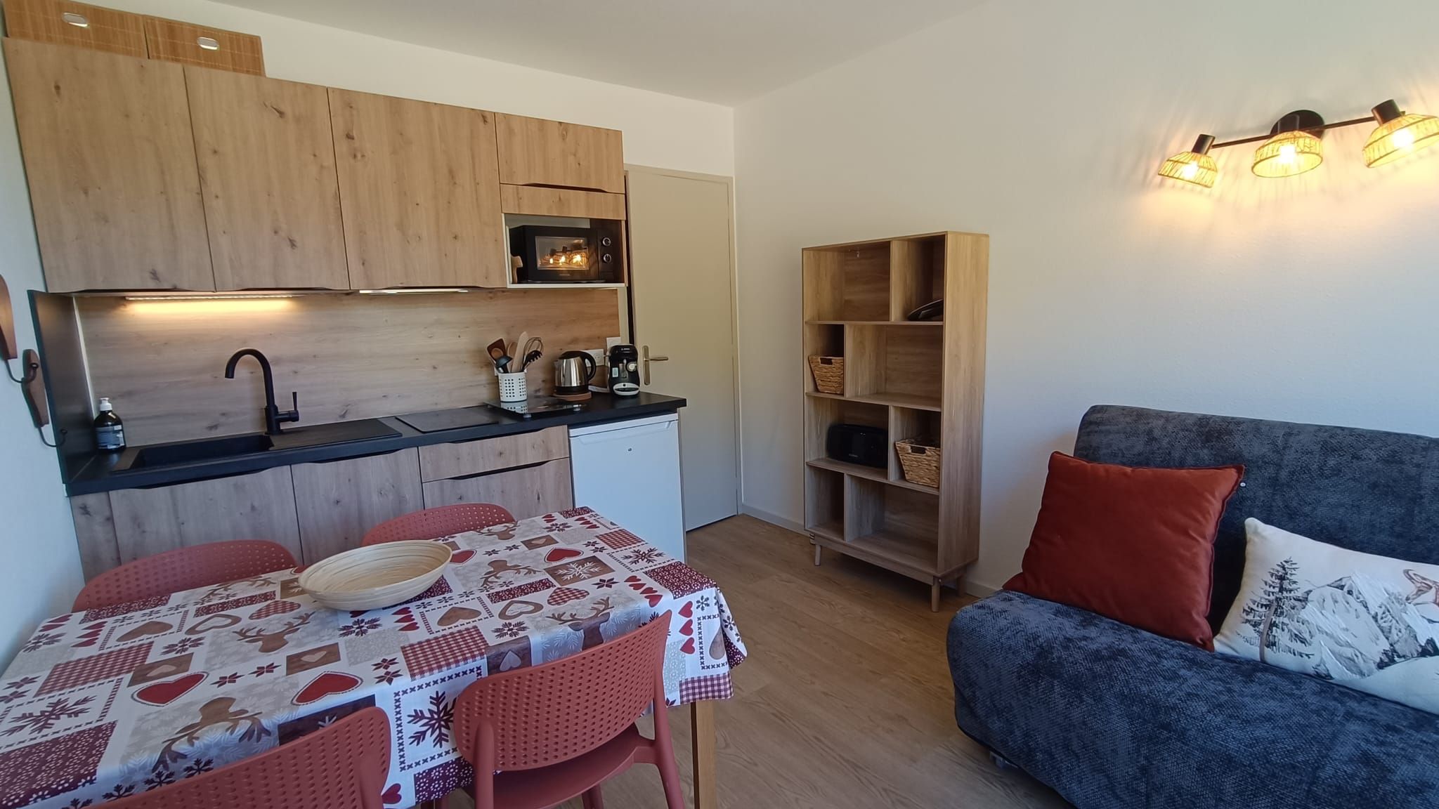 Een kleine keuken met houten kastjes, een eettafel met een feestelijk tafelkleed, en een blauwe bank met rode kussens.