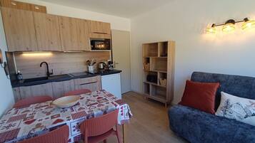 Een kleine keuken met houten kastjes, een eettafel met een feestelijk tafelkleed, en een blauwe bank met rode kussens.