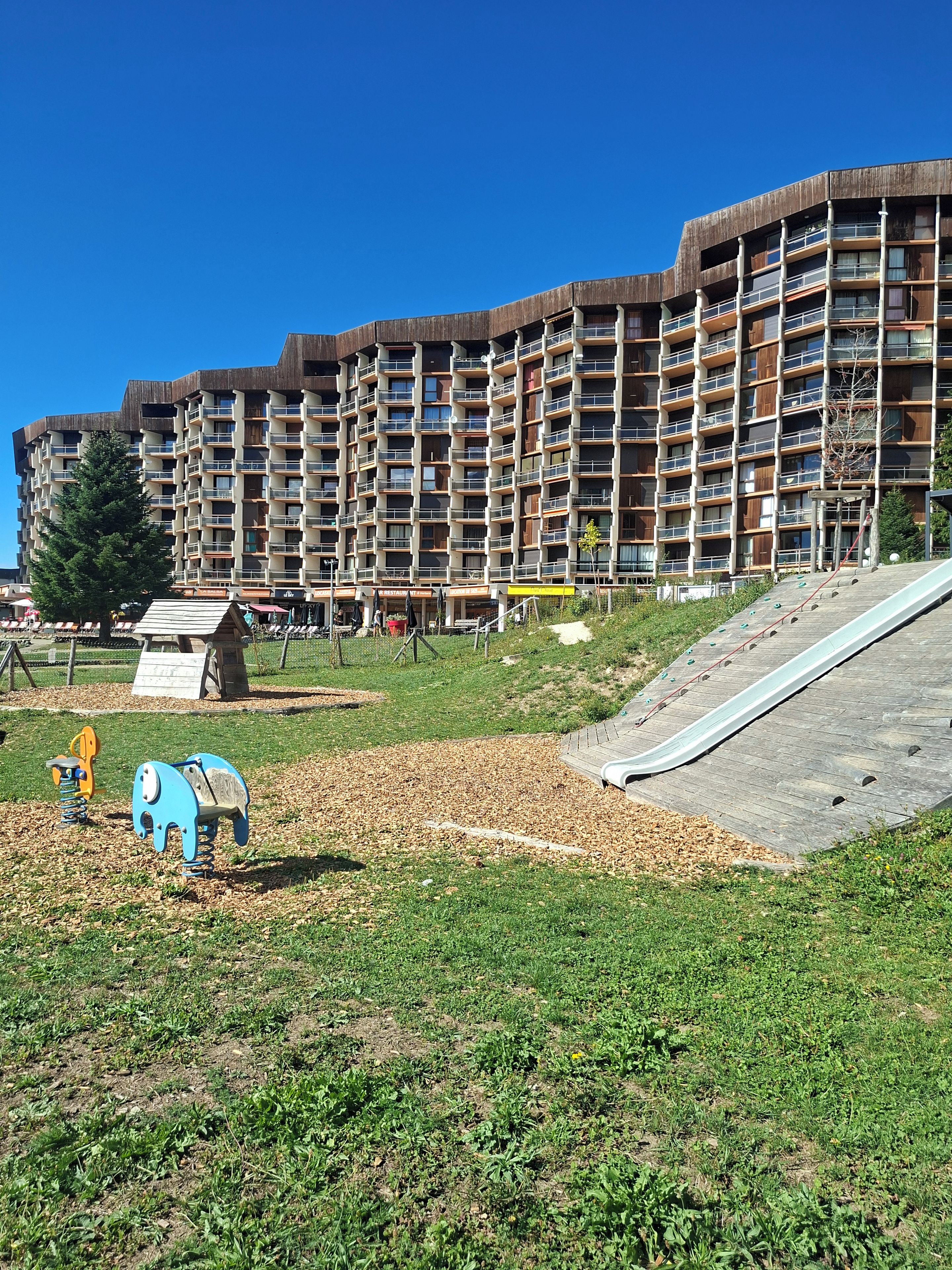 Un edificio residencial de varios pisos con balcones, un parque infantil con tobogán y una zona verde con juegos.