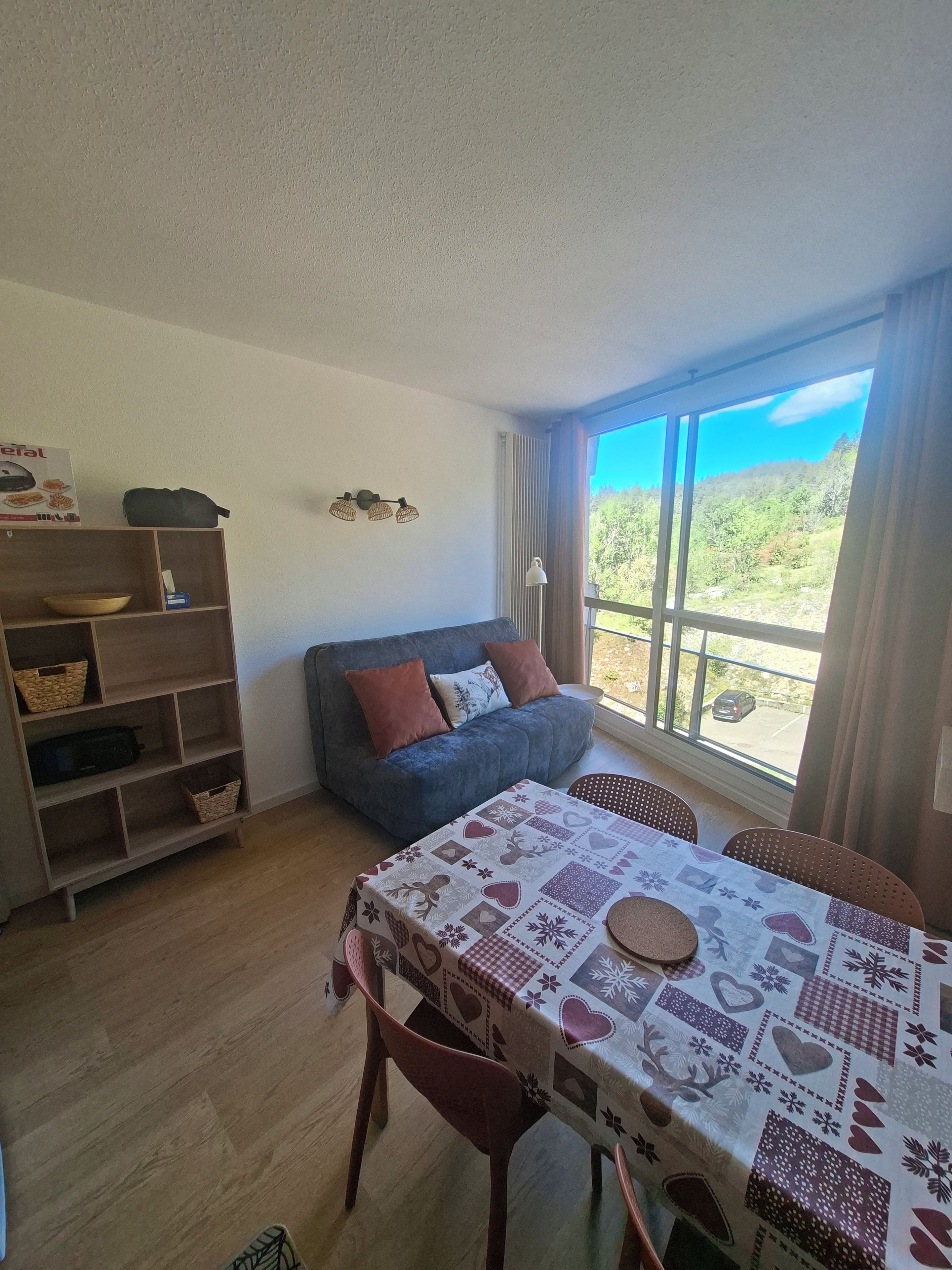 En hyggelig stue med en sofa, et spisebord med stole, en reol og et stort vindue med udsigt over grønne områder.
