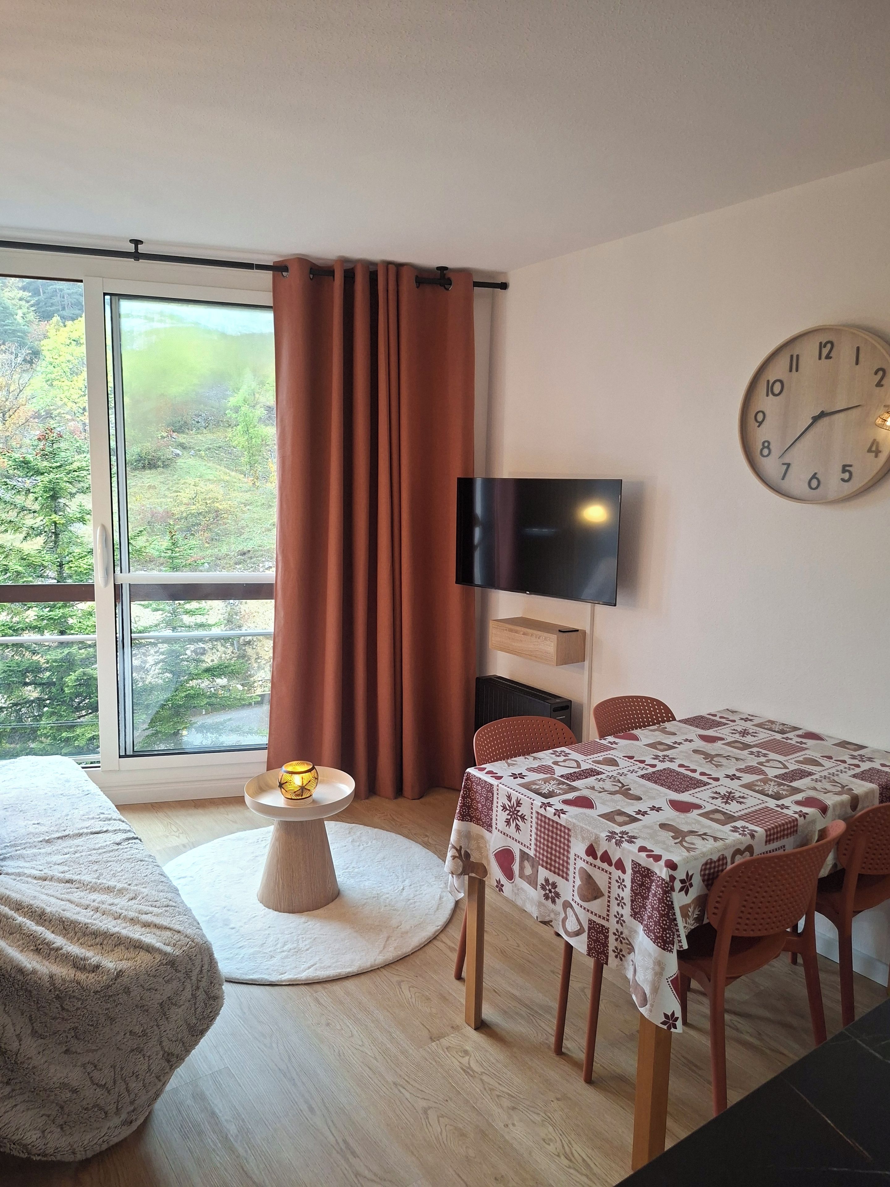 Une pièce comprenant un lit, une table à manger avec des chaises, un téléviseur fixé au mur et une fenêtre avec des rideaux.