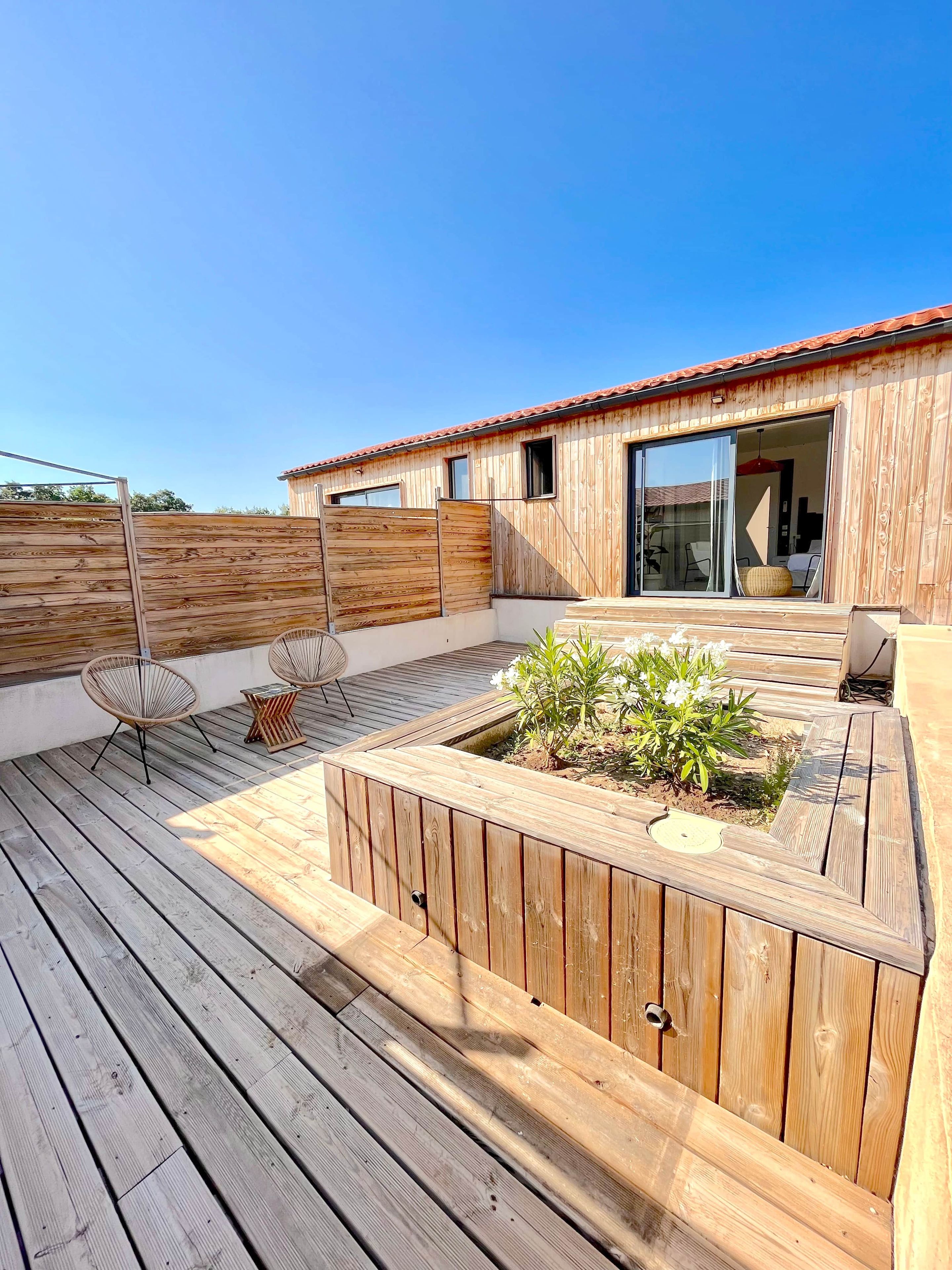 Une terrasse en bois dotée d’un jardin intégré, de deux fauteuils en osier et d’une porte-fenêtre coulissante donnant sur un espace intérieur.