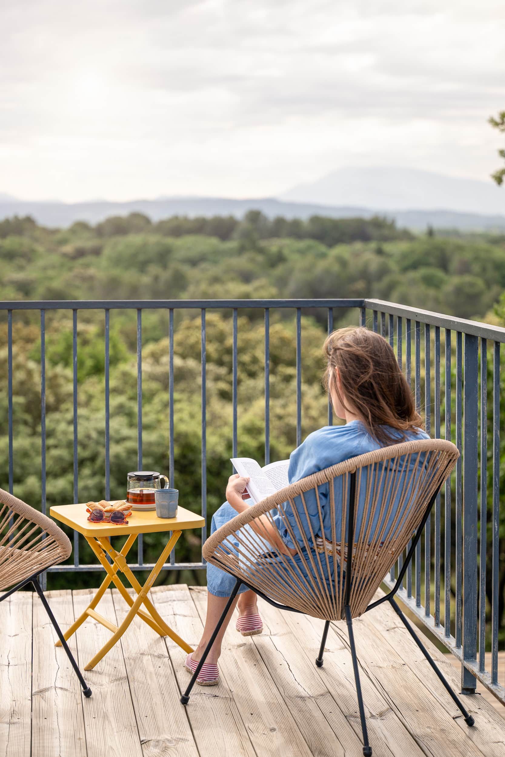 Eine Person sitzt auf einem Korbstuhl auf einer Holzterrasse und liest ein Buch. Ein Tisch nebenan trägt eine Tasse, Gläser und einen Behälter.