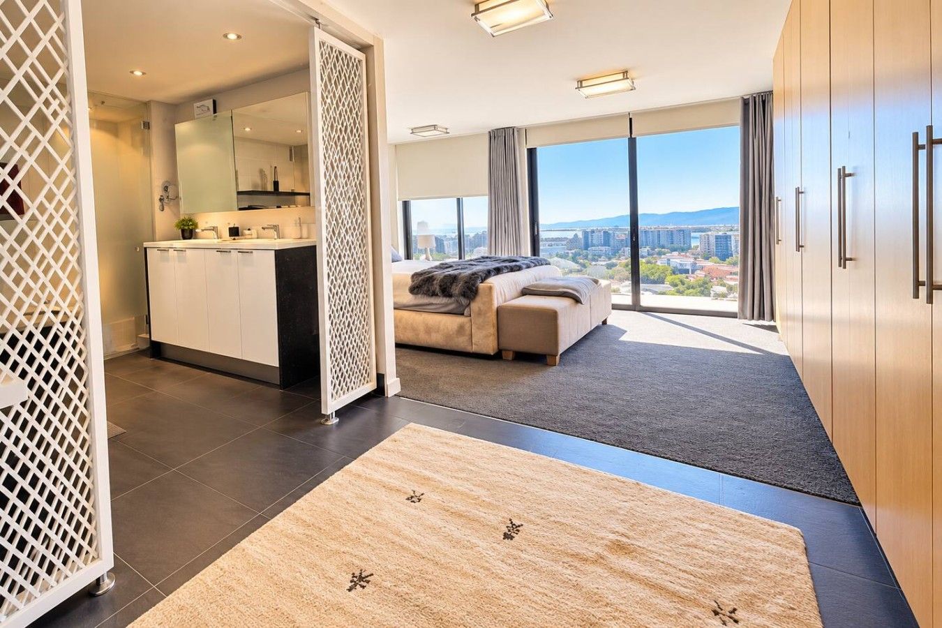 A modern hotel room with a large bed, a separate bathroom, a kitchenette, and a view of the city.