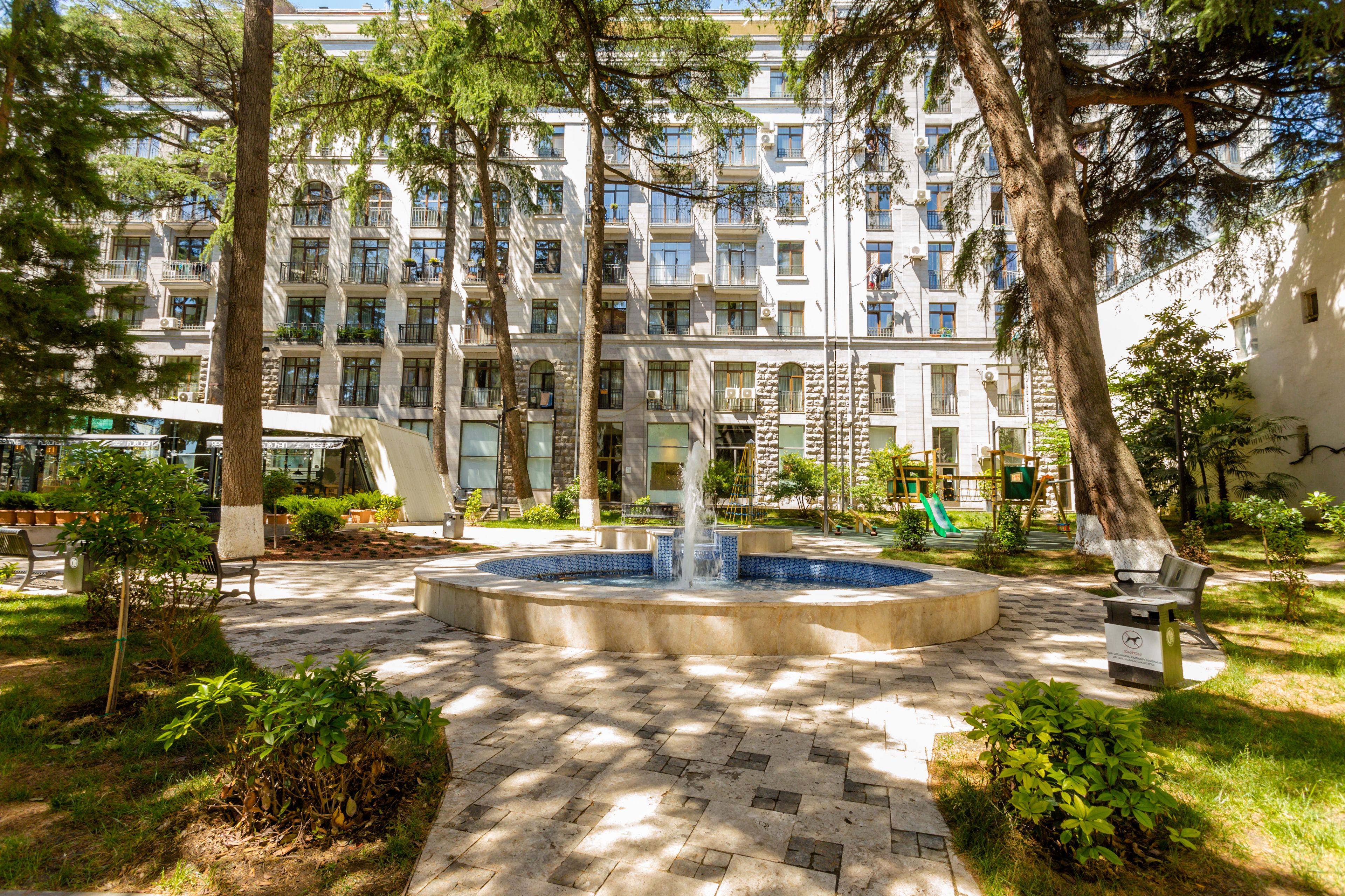 A courtyard with a fountain, trees, and a building with balconies in the background.
