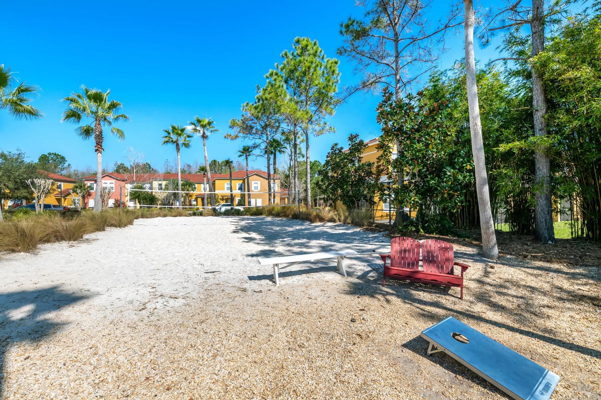 A sandy beach area with a red bench and a lounge chair, surrounded by trees and buildings in the background.