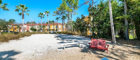 A sandy beach area with a red bench and a lounge chair, surrounded by trees and buildings in the background.