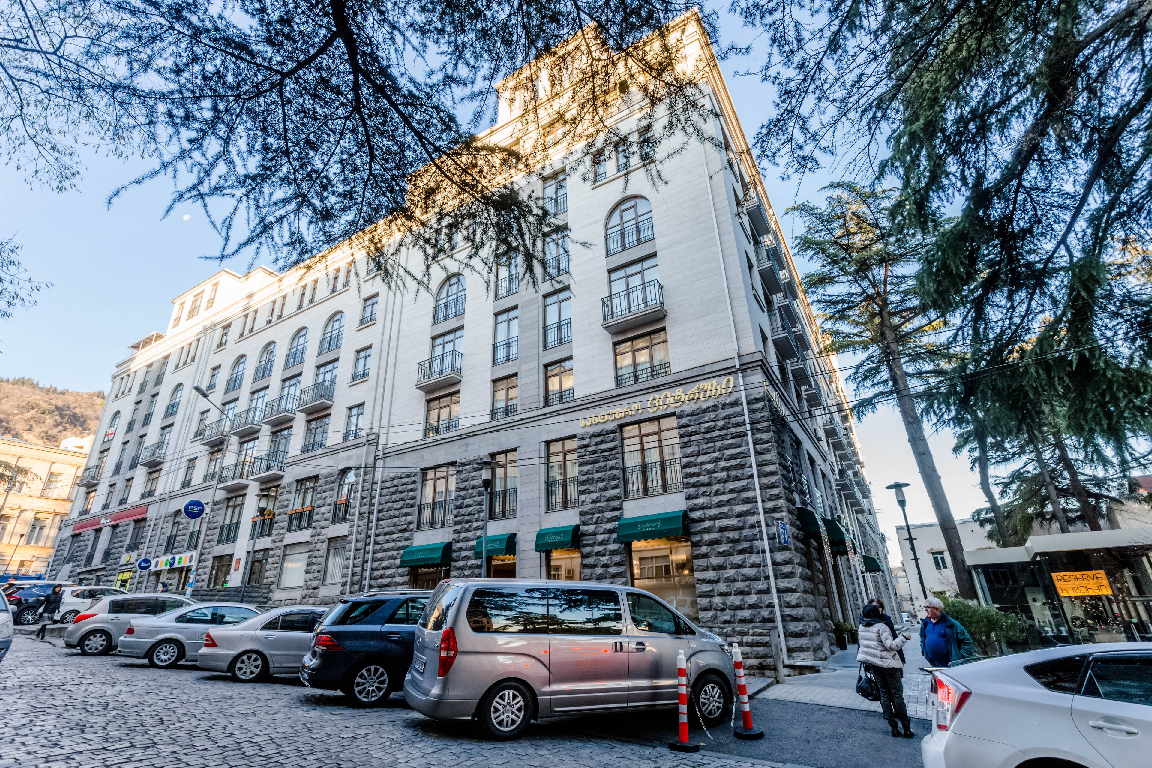 A multi-story hotel with a stone facade, multiple balconies, and a row of parked cars in front.