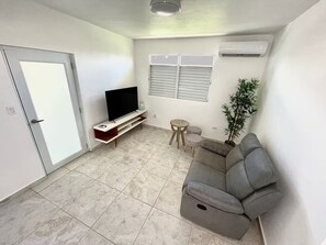 A living room with a grey sofa, a flat-screen TV on a stand, a small round table, a potted plant, and a door with a white frame. - Bucketlist Private Beach Villa Walk Beach-2ndFloor (San Juan)