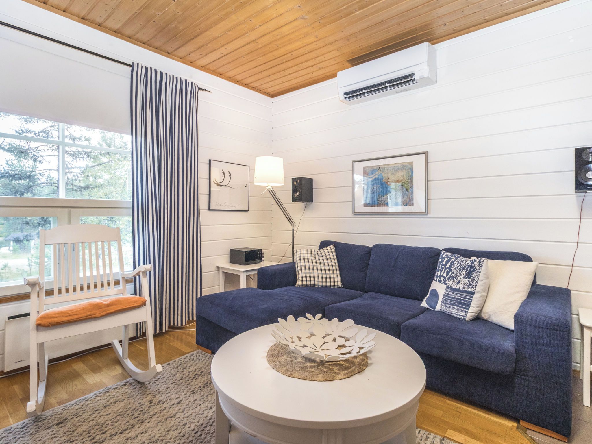 Una sala de estar con un sofá azul, un sillón mecedor blanco, una mesa de centro redonda y una ventana con cortinas.