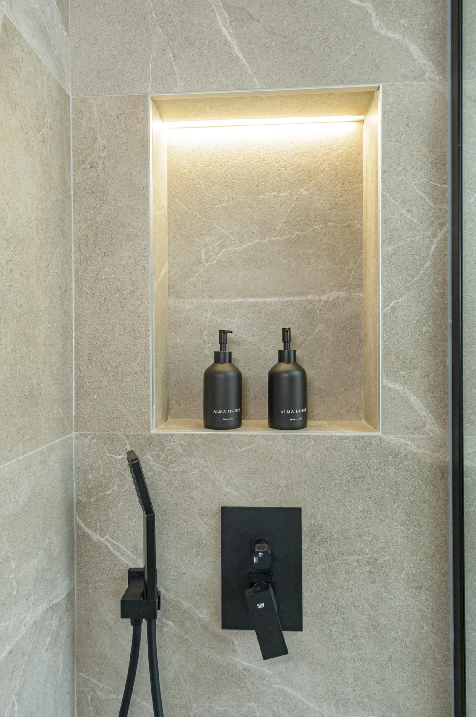 Een badkamer met een ingebouwd schap met twee flessen ALMA HOUSE shampoo en douchegel, en een wandpaneel voor de douchekraan.