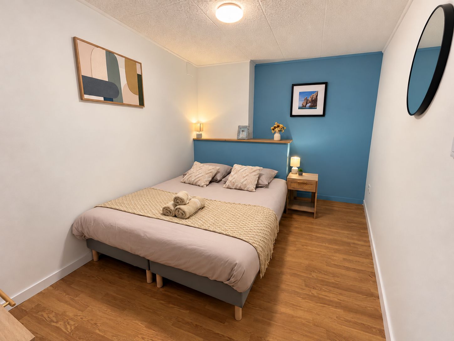 A bedroom with a bed, bedside tables, a wall-mounted mirror, and artwork.