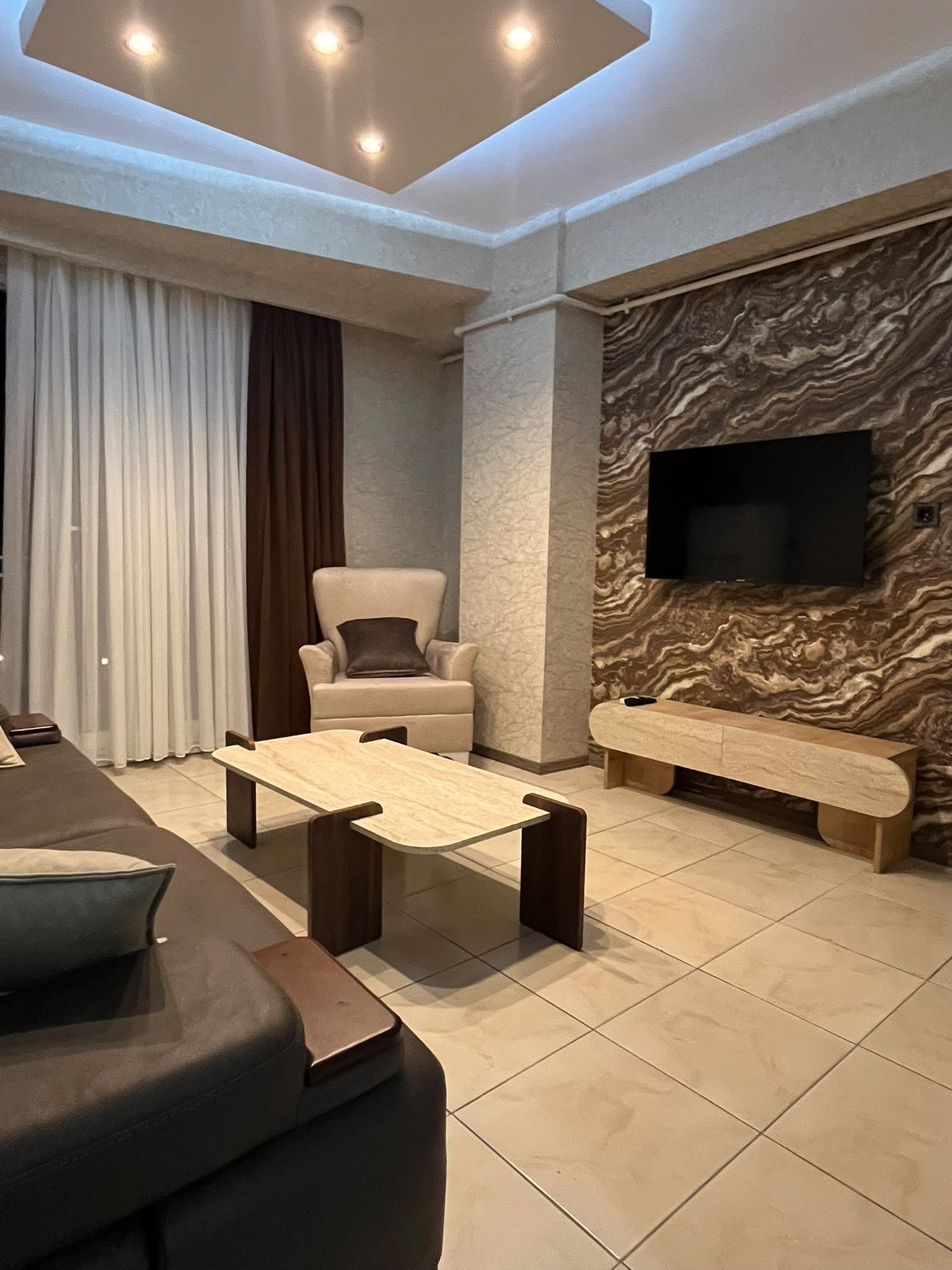 A modern living room with a sofa, a coffee table, a chair, a flat-screen TV mounted on a textured wall, and a large window with curtains.