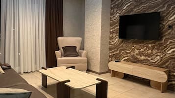 A modern living room with a sofa, a coffee table, a chair, a flat-screen TV mounted on a textured wall, and a large window with curtains.