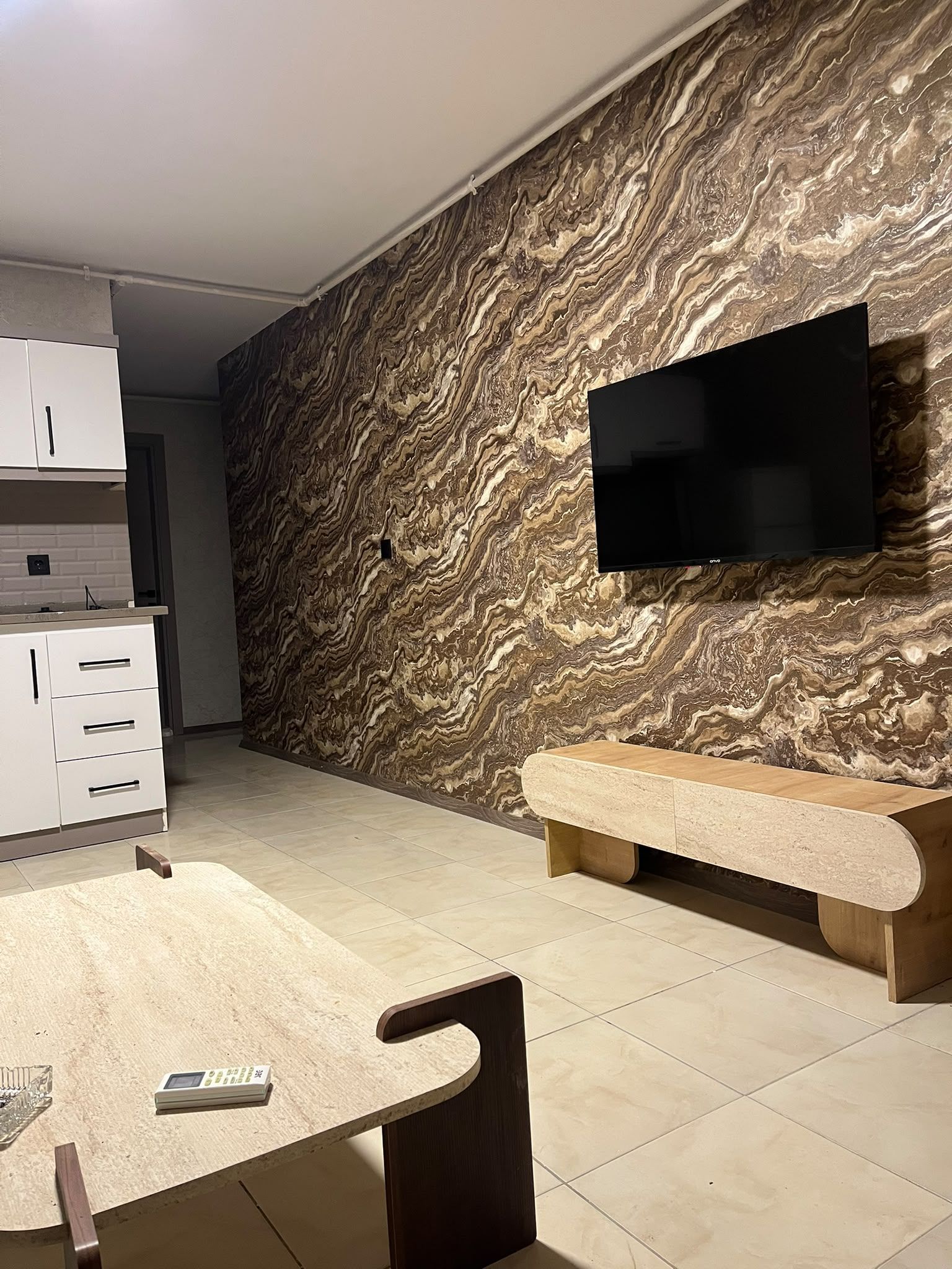Ett modernt kök med en väggbeklädnad av marmor, en platt-TV som är monterad på väggen, en inbyggd bänk och en köksö med en digital våg.
