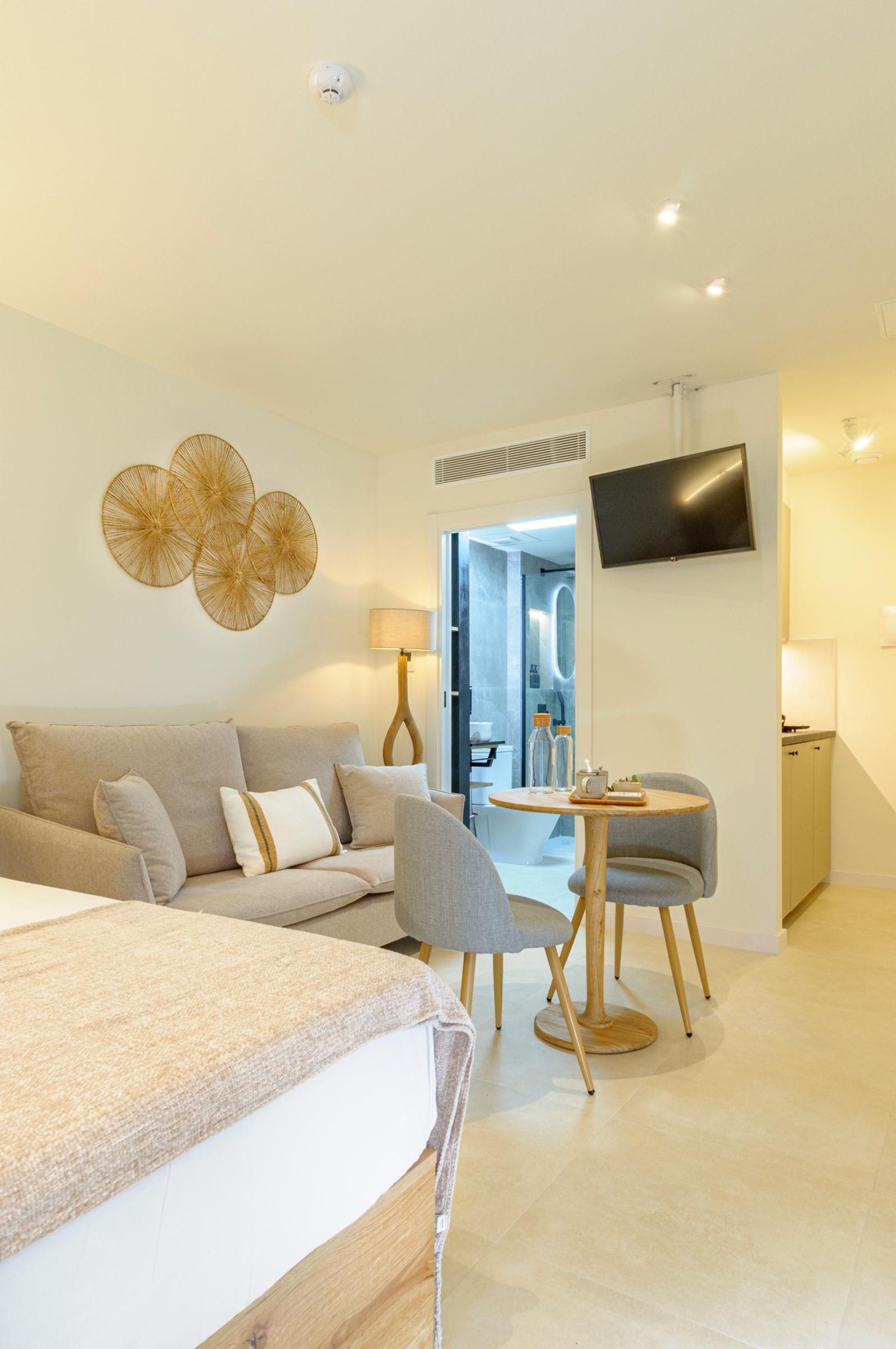 Een moderne hotelkamer met een bed, een bank, een eettafel met stoelen, een aan de muur bevestigde televisie en een badkamer die door een geopende deur zichtbaar is.