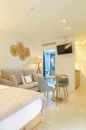 Ein modernes Hotelzimmer mit Bett, Sofa, Esstisch mit Stühlen, einem an der Wand befestigten Fernseher und einem durchgehenden Türspalt sichtbaren Badezimmer.