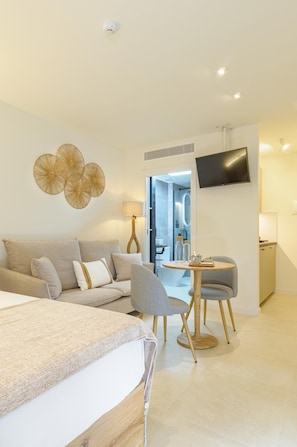 Ein modernes Hotelzimmer mit einem Bett, einem Sofa, einem Esstisch mit Stühlen, einem an der Wand befestigten Fernseher und einem durch eine offene Tür einsehbaren Badezimmer.
